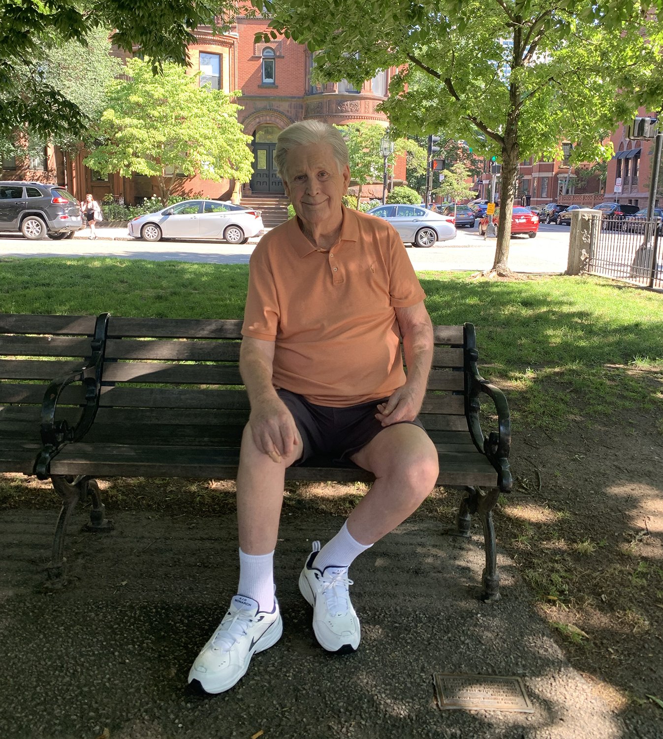 Brian sitting on a park bench under a tree with cars and a red brick building in the background in Boston.