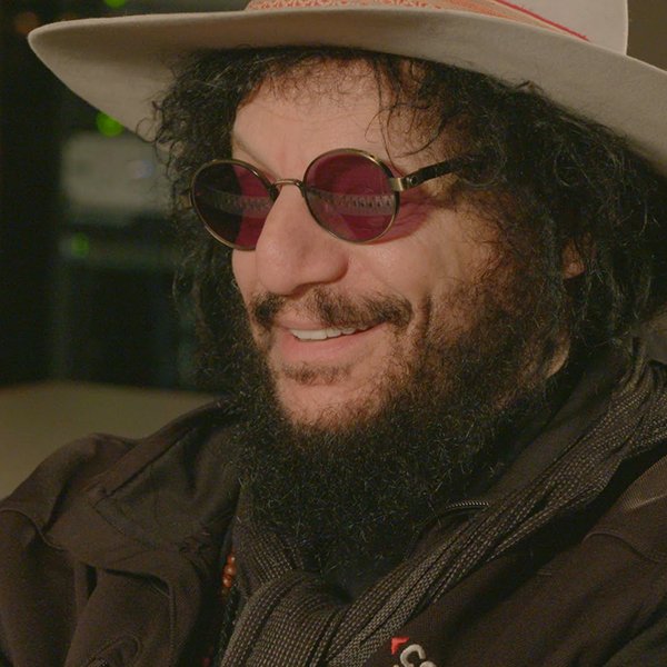 Close-up of a man with curly hair, beard, and sunglasses, wearing a light-colored hat and smiling.