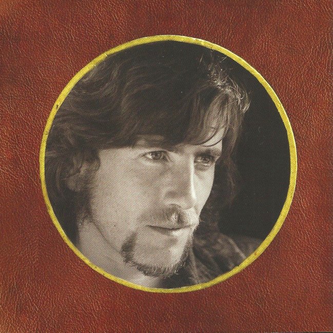 Black and white portrait of a man with medium-length wavy hair, a beard, and a mustache, inside a circular frame with a yellow border on a textured brown background.