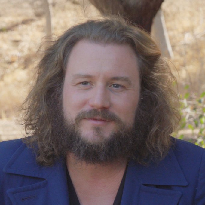 A man with long, curly hair and a beard, wearing a dark blue shirt outdoors with trees and sunlight in the background.