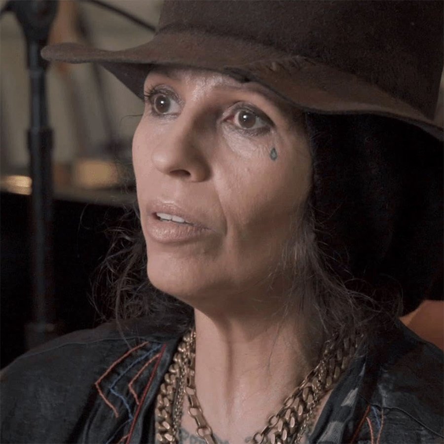 Close-up of a woman with a teardrop tattoo, wearing a brown hat, layered necklaces, and a black leather jacket, looking slightly upwards.